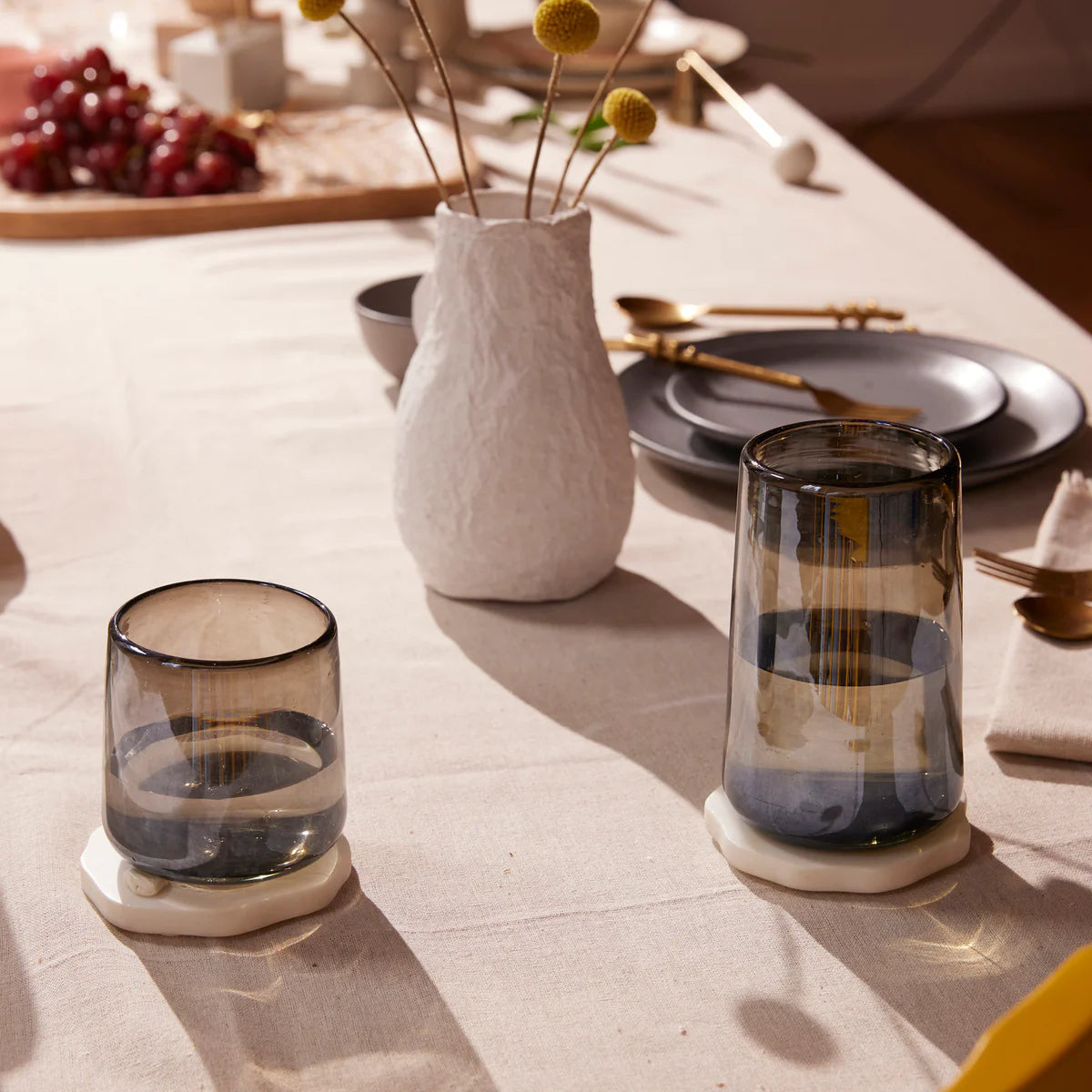Two glass candle holders on a table with a vase and fruit in the background