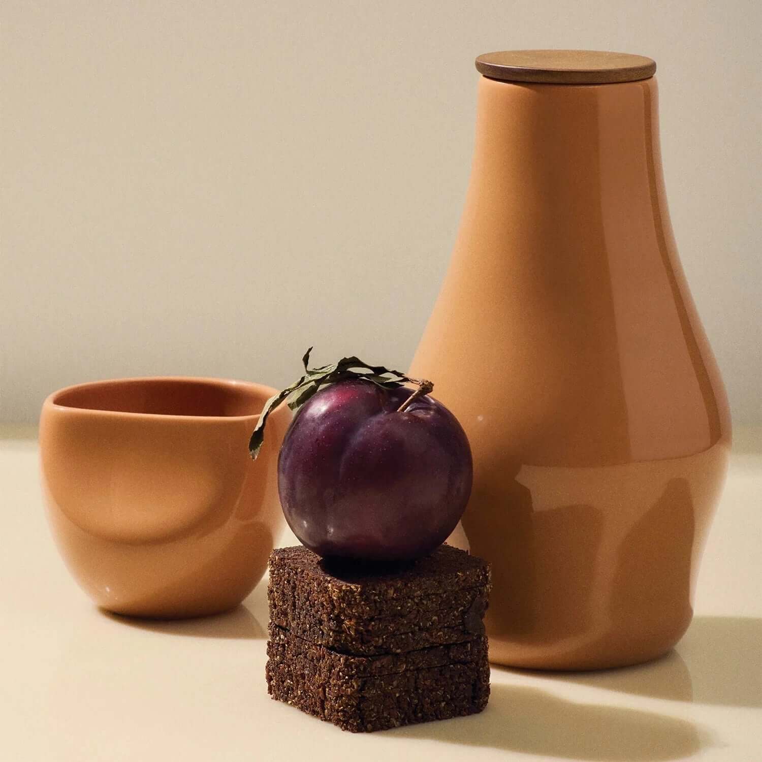 Terracotta pitcher, cup, plum, and dark brown square on a light background